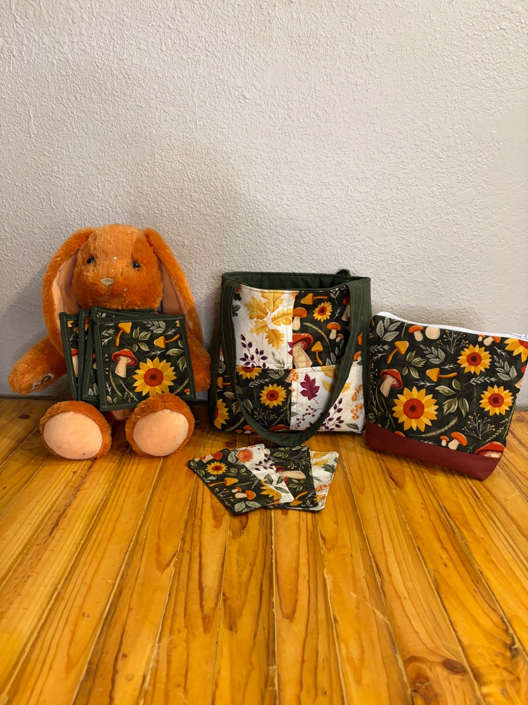 A handmade tote, pouch, coasters, and pot holders in sunflower fabric, arranged on a pine table next to a plush bunny